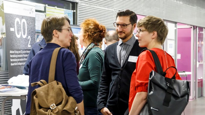 Das Bild zeigt vier Personen, die vor einem Informationsstand stehen, auf dem ein Poster mit der Aufschrift „-CO2“ zu sehen ist. Sie tragen unterschiedliche Kleidung und haben Taschen dabei, was darauf hindeutet, dass sie sich für Informationen am Stand interessieren. Der Stand befindet sich im hellbeleuchteten Foyer der Hochschule Neu-Ulm. 