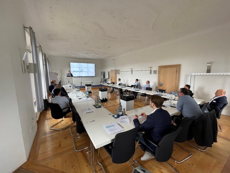 (opens enlarged image) Das Bild zeigt einen Konferenzraum mit Personen, die an Tischen in U-Form sitzen und zur Präsentationswand vorne schauen. Auf der Wand ist eine Folie mit Text und Grafiken zu sehen, die vermutlich im Rahmen einer Präsentation oder eines Meetings dient. Der Raum ist gut beleuchtet und verfügt über dekorative Deckendetails. (opens enlarged image)