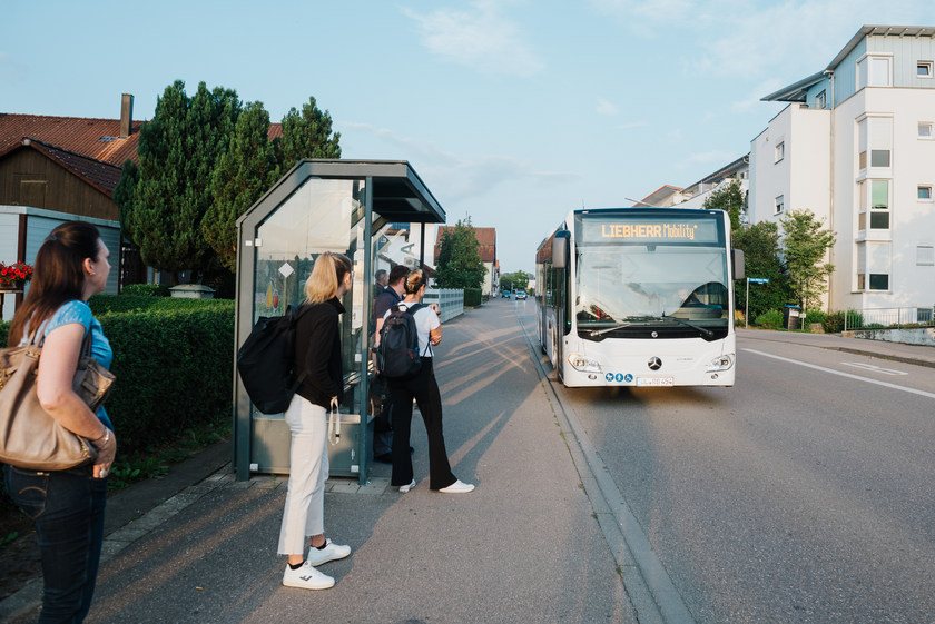 Das Bild zeigt Menschen an einer Bushaltestelle stehen. Der Bus hält neben der Bushaltestelle an.