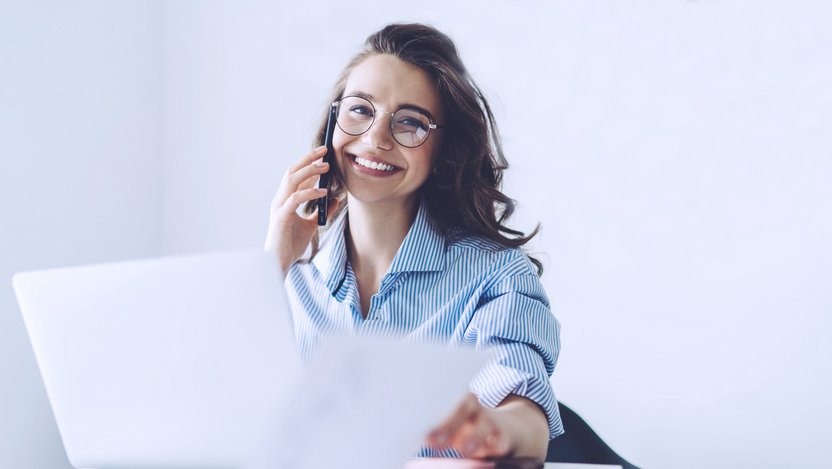 Smiling student in striped blouse sits behind a laptop, leafs through a folder, makes a phone call and looks into the camera