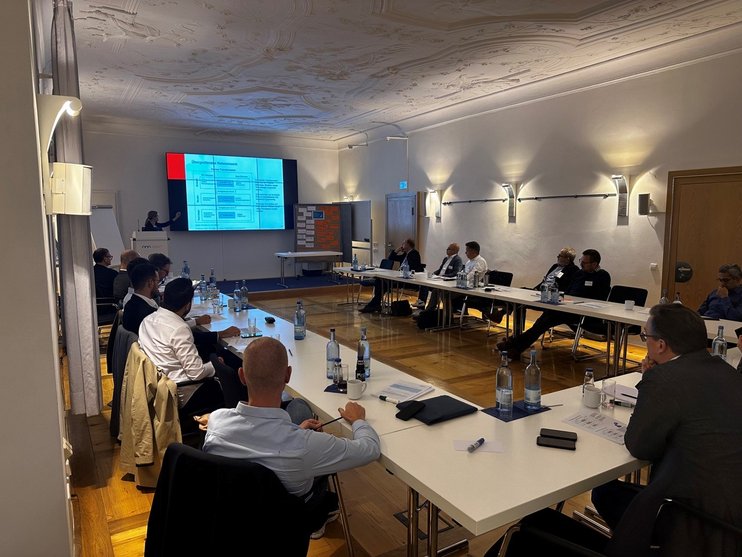 (opens enlarged image) Das Bild zeigt einen Konferenzraum mit Personen, die an Tischen in U-Form sitzen und zur Präsentationswand vorne schauen. Auf der Wand ist eine Folie mit Text und Grafiken zu sehen, die vermutlich im Rahmen einer Präsentation oder eines Meetings dient. Der Raum ist gut beleuchtet und verfügt über dekorative Deckendetails. Die Fenster auf der linken Seite lassen Tageslicht herein. Dieses Setting scheint für Bildungs-, Geschäfts- oder Organisationszwecke relevant zu sein, bei denen Präsentationen oder Besprechungen durchgeführt werden. (opens enlarged image)