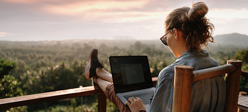 [Translate to English:] Schmuckbild: Frau mit Laptop blickt auf sonnige, hügelige Landschaft