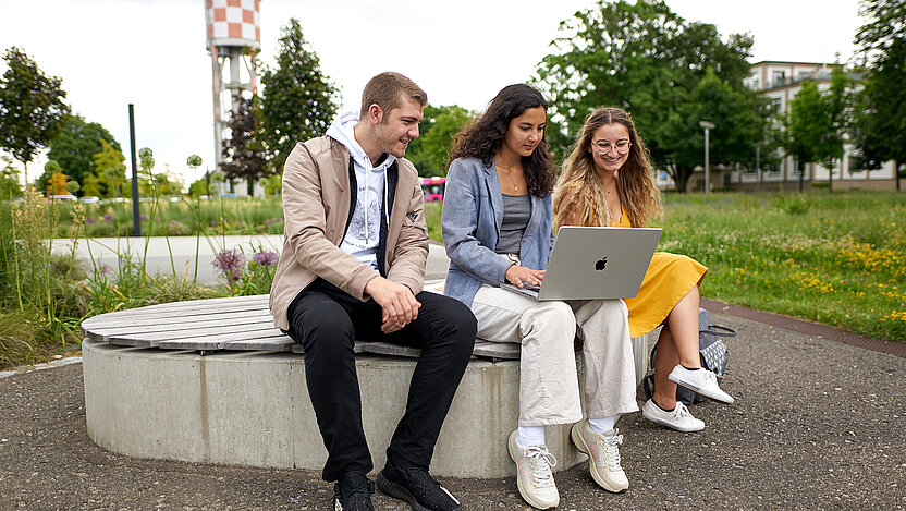 Ein junger Mann und zwei junge Frauen sitzen auf einer Bank vor der Hochschule, schauen in einen Laptop und informieren sich über das Studienangebot