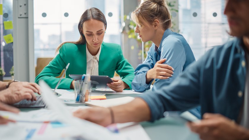 Portrait of Two Creative Colleagues Collaborating and Pointing at Digital Tablet in Office Hub. White Female Designer Discussing Work with Assistant and Choosing Marketing Strategy. Coworkers Working