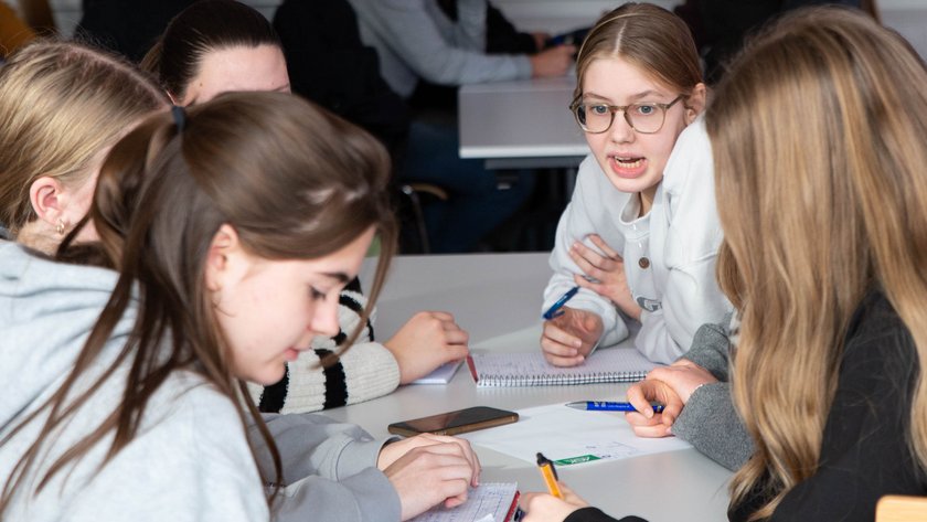 Pupils sit at a table and talk to each other and write on paper. (opens enlarged image)