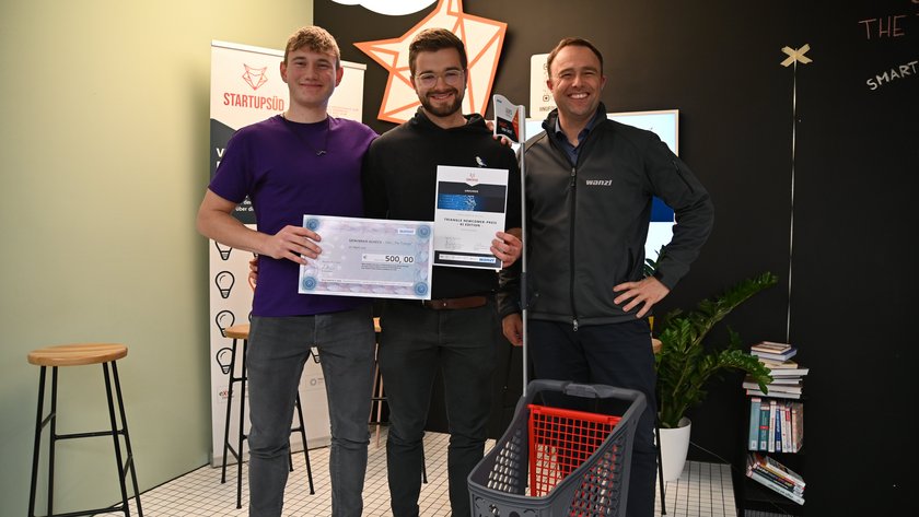 Das Bild zeigt zwei Personen, die einen großen Scheck und eine Urkunde halten. Dies deutet darauf hin, dass sie einen Preis oder eine Auszeichnung gewonnen haben. Eine weitere Person steht daneben und vor dieser Person ist ein Einkaufswagen zu sehen. Es handelt sich um einen Moment der Leistung oder Anerkennung.