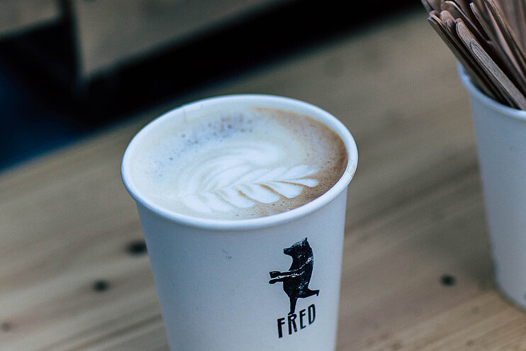Eine Tasse mit dem Kaffee-Fred-Logo, dem braunen Bären. In der Tasse ist ein Cappucino angerichtet. Die Milchhaube ist mit einem Blattmuster verziert. (öffnet Vergrößerung des Bildes)