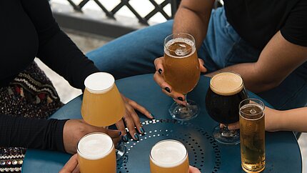 [Translate to English:] Das Bild zeig einen Bildausschnitt von sechs Personen die gemeinsam an einem Tisch sitzen und Bier trinken. Der Ausschnitt zeigt nur den Tisch mit Bier und Händen der Personen.
