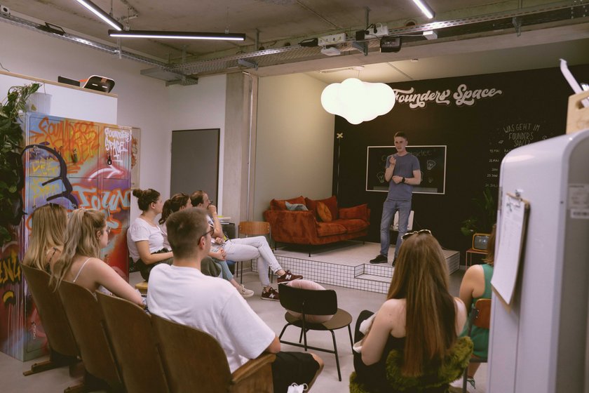 Das Bild zeigt den HNU Founders Space. Hier präsentiert eine Person vor einer Gruppe von Menschen. Diese sitzen auf Stühlen oder Sofas. Im Hintergrund der präsentierenden Person ist eine Wandtafel mit der Aufschrift „Founders Space“ zu erkennen.  (öffnet Vergrößerung des Bildes)