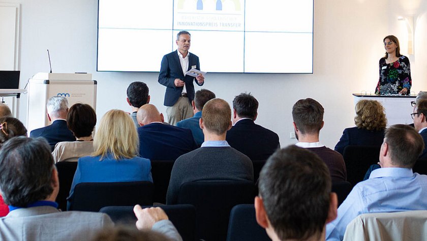 Das Bild zeigt eine männliche Person, die vor einem Publikum steht. Das Publikum ist nur von hinten zu sehen. Der Präsentator steht vor einem weißen Bildschirm und spricht zu dem Publikum. 