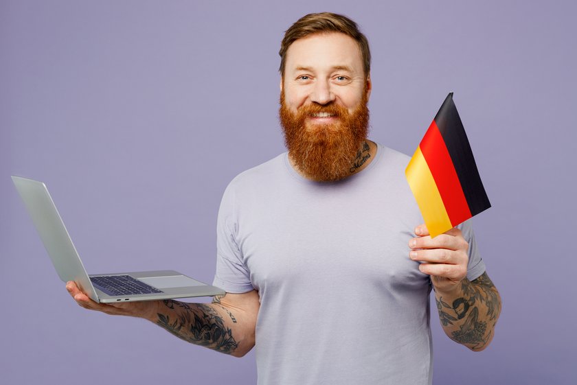 Young redhead bearded IT man he wear violet t-shirt casual clothes hold german flag use work on laptop pc computer isolated on plain pastel light purple background studio portrait. Lifestyle concept. Young redhead bearded IT man he wear violet t-shirt casual clothes hold german flag use work on laptop pc computer isolated on plain pastel light purple background studio portrait. Lifestyle concept.