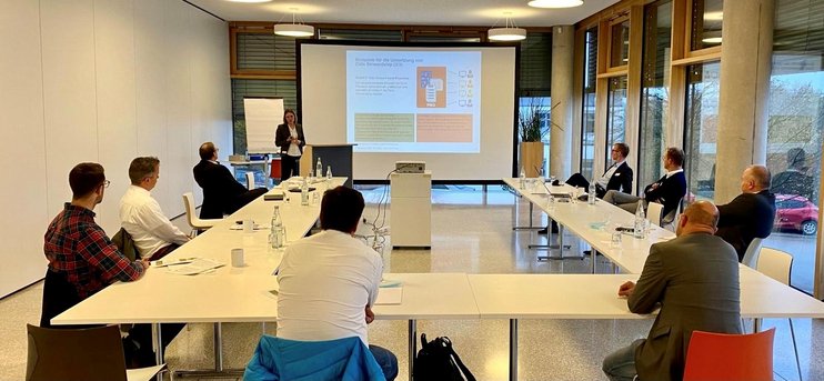 (opens enlarged image) Das Bild zeigt einen Klassenraum oder einen Konferenzraum mit mehreren Personen, die an Tischen in U-Form sitzen und zur Präsentationswand vorne schauen. Auf der Wand ist eine Folie mit grafischen Elementen und Text zu sehen, aber der genaue Inhalt ist aus der Ferne nicht erkennbar. Der Raum verfügt über große Fenster auf einer Seite, die Tageslicht hereinlassen, und auf den Tischen liegen verschiedene persönliche Gegenstände wie Notizbücher und Stifte, was auf eine Bildungs- oder berufliche Veranstaltung hindeutet. (opens enlarged image)