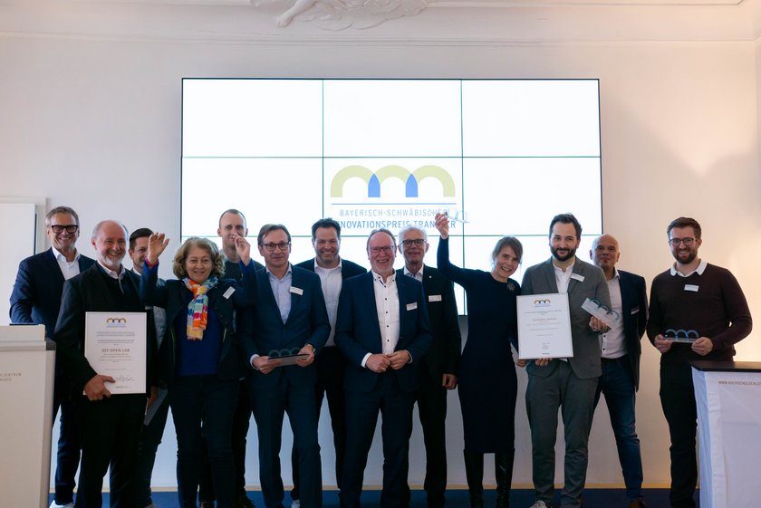 Das Bild zeigt eine Gruppe von Menschen vor einer Wand stehen. Einige der Menschen halten eine Auszeichnung in den Händen. Alle Personen sehen glücklich aus. Auf der weißen Wand im Hintergrund ist ein Bildschirm zu erkennen, auf dem das Logo des "Bayerisch-Schwäbischen Innovationspreis Transfer" zu erkennen ist.