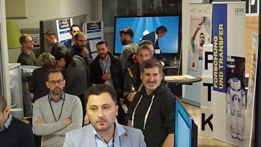 Das Bild zeigt Personen bei einer Veranstaltung in einem hellbeleuchteten Innenraum. Einige der Personen unterhalten sich miteinander. Im Hintergrund ist der Aufsteller "PTK" und "Forschung und Transfer" der Hochschule Neu-Ulm zu erkennen. 