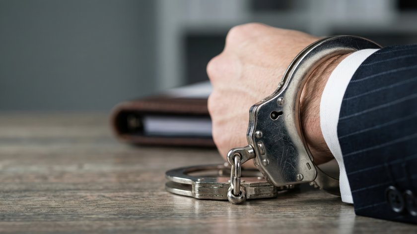Handcuffed Wrist in Business Suit on Desk with Folder Symbolizing White Collar Crime Concept (öffnet Vergrößerung des Bildes) (öffnet Vergrößerung des Bildes)